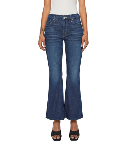A person stands against a white background, hands relaxed by their sides, wearing the Lil’ Weekender mid rise dark blue denim flare jeans by Mother, styled with a white top and black open-toe heels.