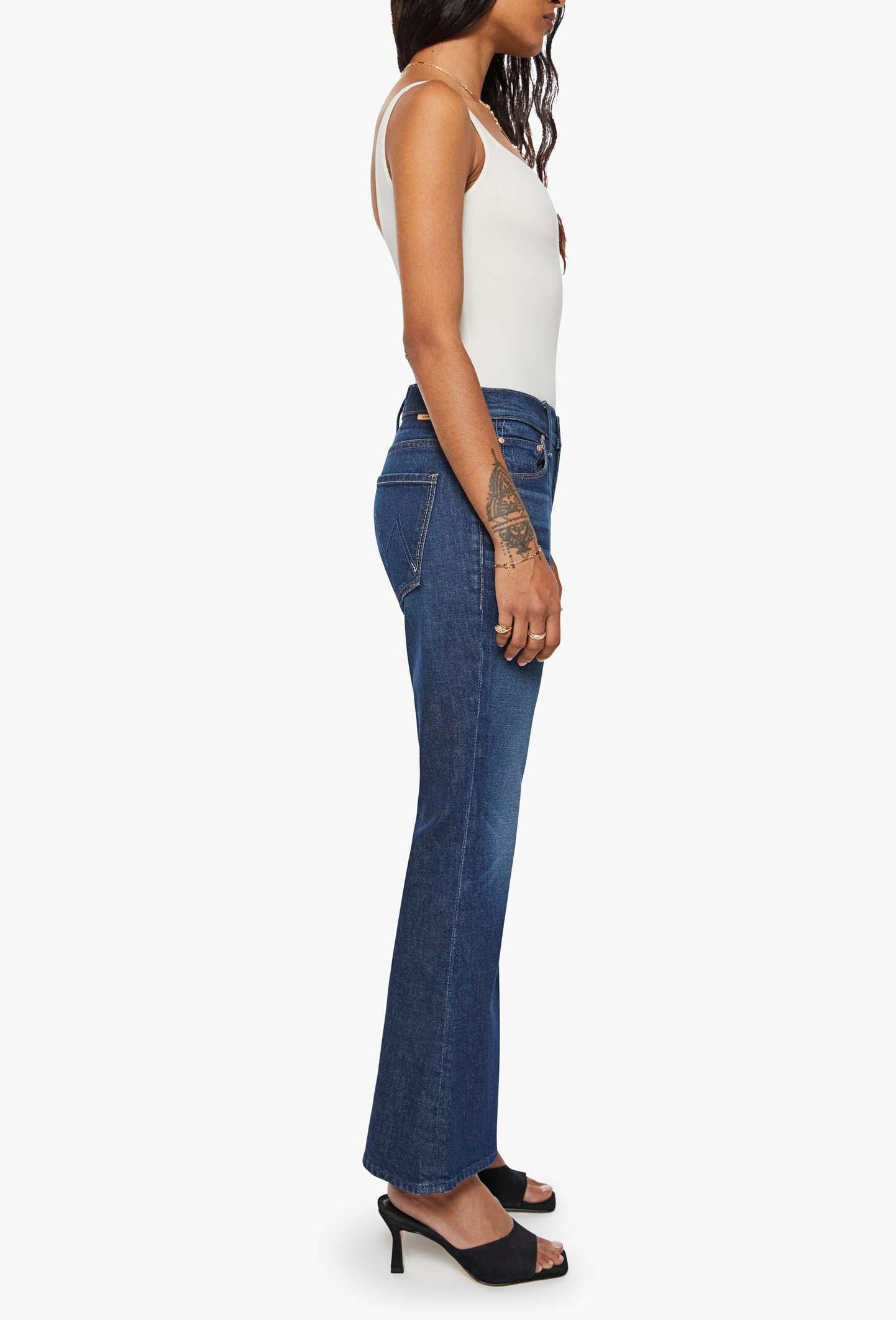 A woman standing in profile wears the Lil’ Weekender dark blue denim flare jeans by Mother with a white tank top and black heels against a plain white background.