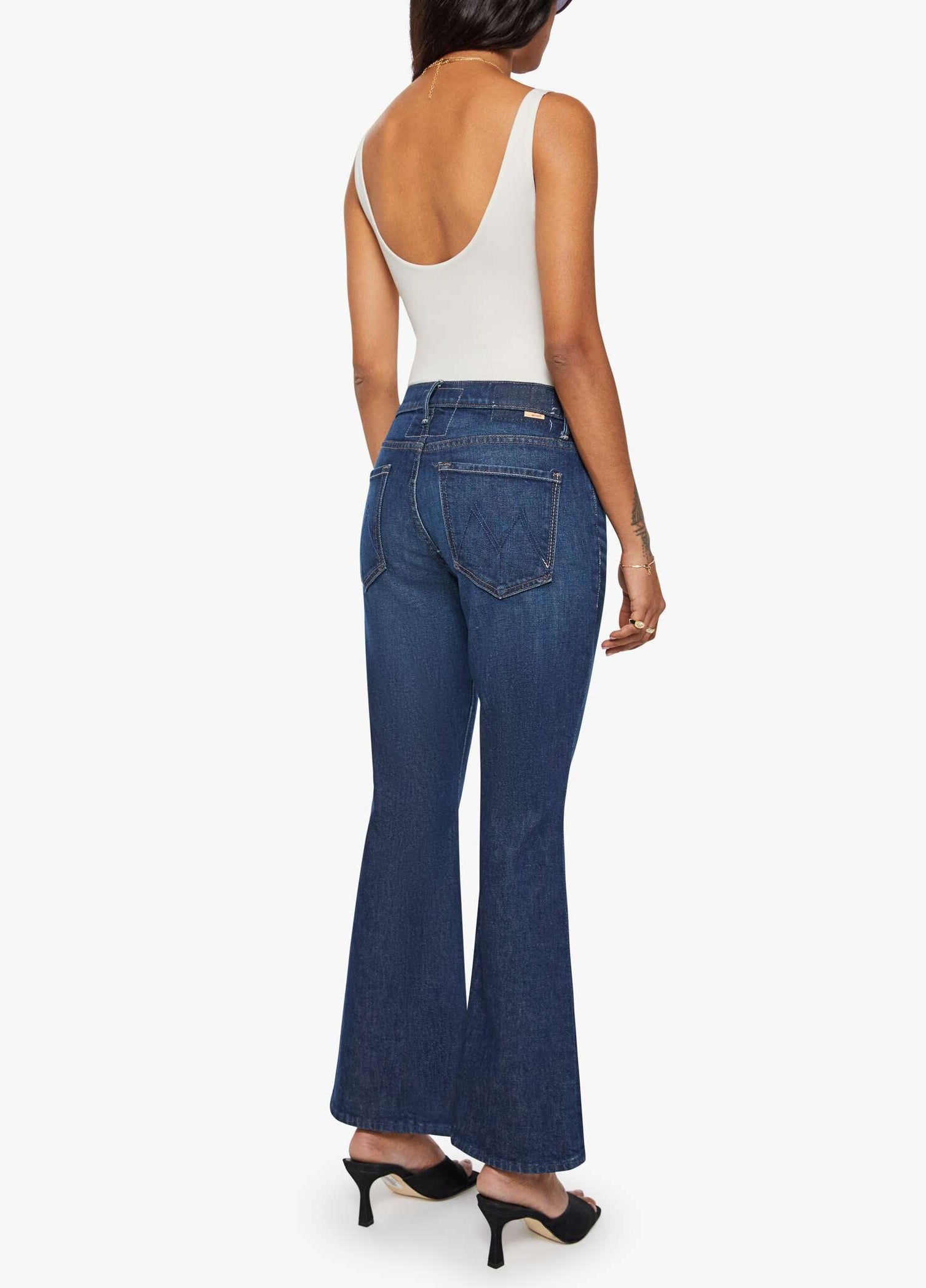 A woman stands facing away, wearing the Lil’ Weekender mid rise dark blue denim flare jeans by Mother, styled with a white sleeveless top and black heeled sandals against a plain white background.