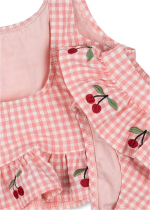 Close-up of the Girls' Soline Bikini by Konges Slojd, featuring pink gingham seersucker fabric with frill details and embroidered red cherries with green leaves.