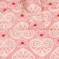 The Konges Slojd Lacy Pink Doll Bag features a pink fabric with white lace-like heart designs, small red hearts, soft shoulder straps, and two handles for easy carrying.