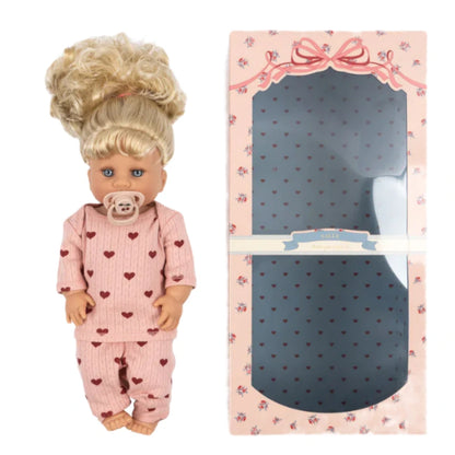 The Konges Slojd Sally Doll, with blonde hair in pink pajamas decorated with red hearts and a pacifier, stands beside an empty pink and blue patterned box.