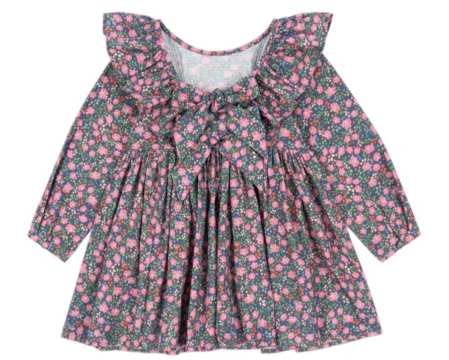 The Konges Slojd Girls' Cianna Bow Dress is a long-sleeve organic cotton dress featuring a ruffled collar, chest bow, and pink floral pattern on a dark background.