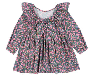 The Konges Slojd Girls' Cianna Bow Dress is a long-sleeve organic cotton dress featuring a ruffled collar, chest bow, and pink floral pattern on a dark background.