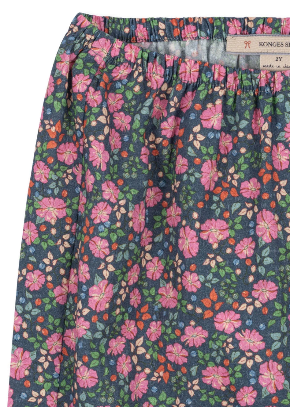A close-up of the Girls' Cianna Pants by Konges Slojd, featuring a blue background with pink, orange, and green floral patterns. The gathered elastic waist ensures comfort, and a partially visible interior label is shown.