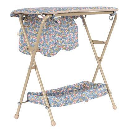 The Konges Slojd Doll Changing Table features a beige metal frame, floral-patterned cover, storage pockets, and a lower shelf for convenience.