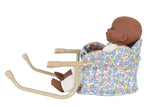 A dark-skinned baby doll seated in the Konges Slojd Doll Table Chair, a floral-patterned, quilted cotton chair with scallop edge and beige metal handles, facing to the right.