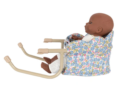 A dark-skinned baby doll seated in the Konges Slojd Doll Table Chair, a floral-patterned, quilted cotton chair with scallop edge and beige metal handles, facing to the right.