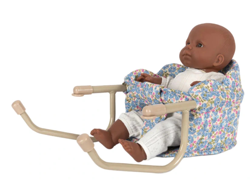 The Konges Slojd Doll Table Chair seats a baby doll in a white outfit, surrounded by a floral print and supported by an iron frame with quilted cotton padding for comfort.