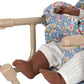 A close-up of a dark-skinned baby doll in white pants and a floral dress seated in a cream-colored Konges Slojd Doll Table Chair with an iron frame.