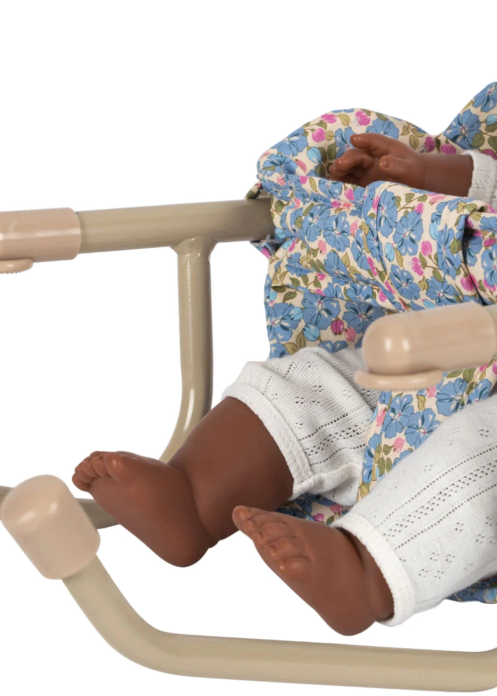 A close-up of a dark-skinned baby doll in white pants and a floral dress seated in a cream-colored Konges Slojd Doll Table Chair with an iron frame.