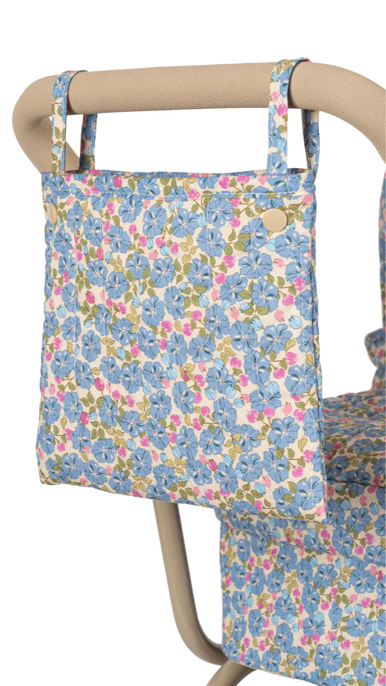 The Konges Slojd Minnie Doll Pram, adorned with a floral-patterned fabric in blue, pink, and green, hangs from a beige chair armrest—perfect for decorating any vintage doll pram or foldable doll stroller.