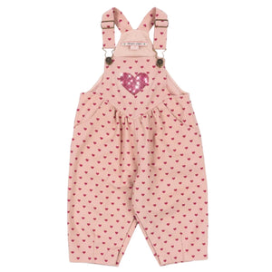 Konges Slojd Baby Girls’ and Girls’ Nola Sequin Overalls feature pink fabric with red heart patterns, a large sequined chest heart, adjustable shoulder straps, and logo buttons at the sides.