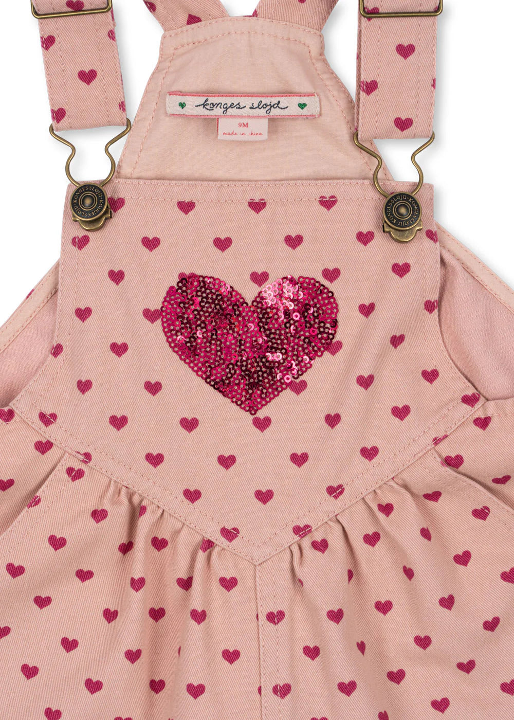The Konges Slojd Baby Girls' and Girls' Nola Sequin Overalls feature a pink heart print, a sparkly sequin heart on the chest, adjustable brass buckle straps, and chic logo buttons for extra style.