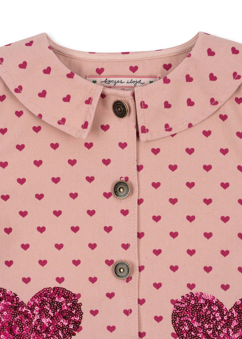 Close-up of the Konges Slojd Baby Girls' and Girls' Nola Sequin Shirt Jacket, a pink cotton twill design with a rounded collar, pink buttons, and small dark pink heart pattern. The label reads "konges sløjd.