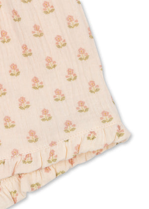 Light pink fabric adorned with a repeating pattern of small pink flowers and green leaves, finished with a ruffled edge at the bottom right corner—ideal for crafting airy and comfortable shorts like the Konges Slojd Baby Girls’ and Girls’ Coco Shorts, which feature an elastic waist for added comfort.