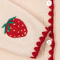 Close-up of Konges Slojd's Girls' Collette Cardigan in beige, featuring red zigzag trim, mother of pearl buttons, and an embroidered red strawberry with green leaves on the sleeve.