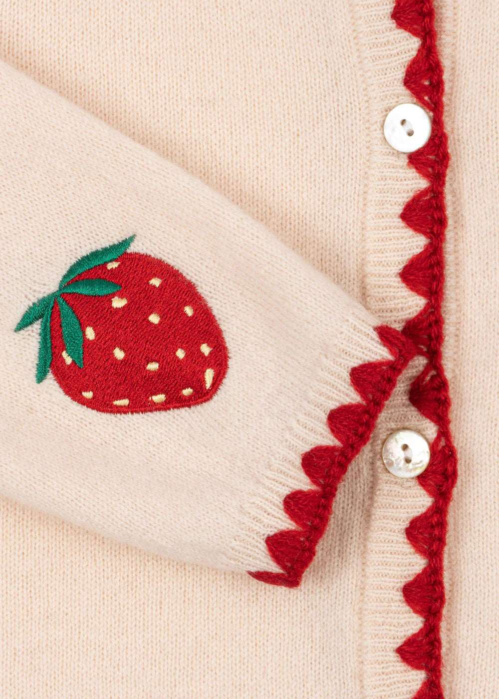 Close-up of Konges Slojd's Girls' Collette Cardigan in beige, featuring red zigzag trim, mother of pearl buttons, and an embroidered red strawberry with green leaves on the sleeve.