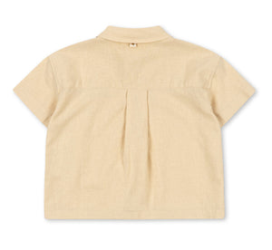The Konges Slojd Baby Boys’ and Boys’ Franzen Short Sleeve Linen Shirt is pictured from the back, showcasing a beige linen-blend design with short sleeves, a collared neckline, central pleat, and a small loop beneath the collar. This breathable shirt delivers comfort and effortless style for everyday wear.