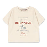 The Konges Slojd Baby Boys’ Itty Tee is a beige short-sleeve top with a boxy fit, featuring the phrase “Just you wait. This is the beginning of something great,” and a small football graphic in the center.