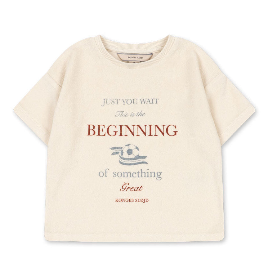 The Konges Slojd Baby Boys’ Itty Tee is a beige short-sleeve top with a boxy fit, featuring the phrase “Just you wait. This is the beginning of something great,” and a small football graphic in the center.