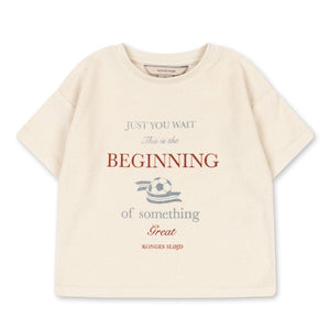 The Konges Slojd Baby Boys’ Itty Tee is a beige short-sleeve top with a boxy fit, featuring the phrase “Just you wait. This is the beginning of something great,” and a small football graphic in the center.