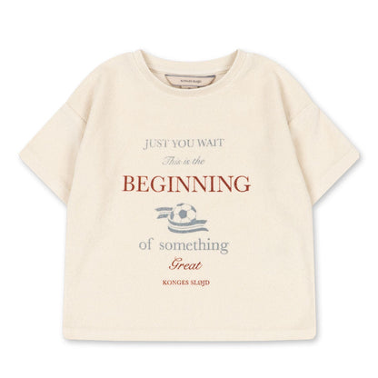 The Konges Slojd Baby Boys’ Itty Tee is a beige short-sleeve top with a boxy fit, featuring the phrase “Just you wait. This is the beginning of something great,” and a small football graphic in the center.