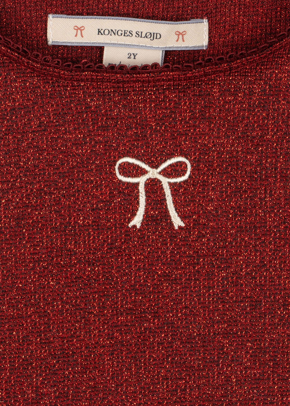 Close-up of a Girls’ Roli Blouse in dark red shimmering glitter with a white embroidered bow and a Konges Slojd brand label at the neckline indicating size 2Y.