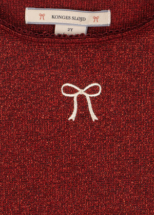 Close-up of a Girls’ Roli Blouse in dark red shimmering glitter with a white embroidered bow and a Konges Slojd brand label at the neckline indicating size 2Y.