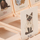 A close-up of the Who Am I Game by Konges Slojd, showing illustrated animal cards and a cat named Kiki in a polka dot scarf.
