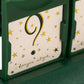 A green game piece holds a card with a bold question mark, surrounded by gold stars and cursive script. Who Am I Game by Konges Slojd is perfect for family guessing fun!.