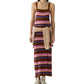A woman wears the Palazzo Dress by Guest in Residence, a sleeveless striped linen knit style in brown, pink, and blue, paired with a sweater tied at the waist and beige sneakers.