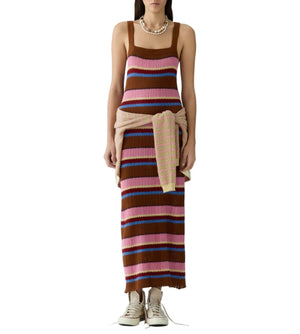 A woman wears the Palazzo Dress by Guest in Residence, a sleeveless striped linen knit style in brown, pink, and blue, paired with a sweater tied at the waist and beige sneakers.