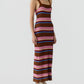 Someone stands against a plain background wearing the Guest in Residence Palazzo Dress, a sleeveless, ankle-length linen knit with brown, pink, purple and red stripes and a ribbed texture, paired with white sneakers and a chunky necklace.