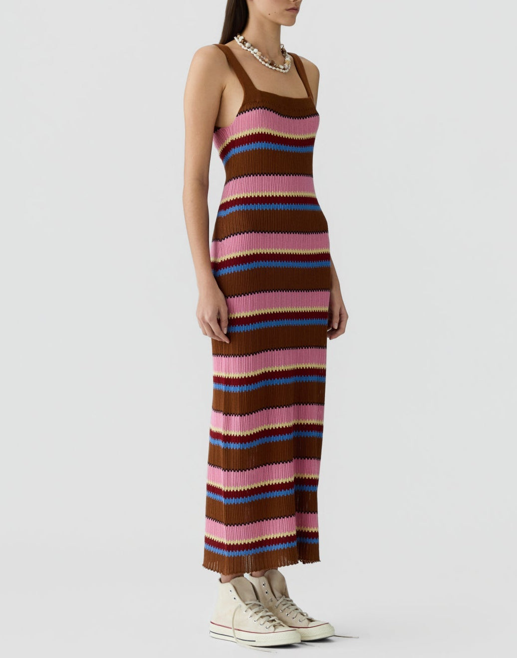 Someone stands against a plain background wearing the Guest in Residence Palazzo Dress, a sleeveless, ankle-length linen knit with brown, pink, purple and red stripes and a ribbed texture, paired with white sneakers and a chunky necklace.