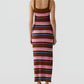 A woman stands facing away, wearing the Guest in Residence Palazzo Dress—a sleeveless, ankle-length silk knit with horizontal stripes in brown, pink, purple, and blue tones—and pairs it with white shoes.