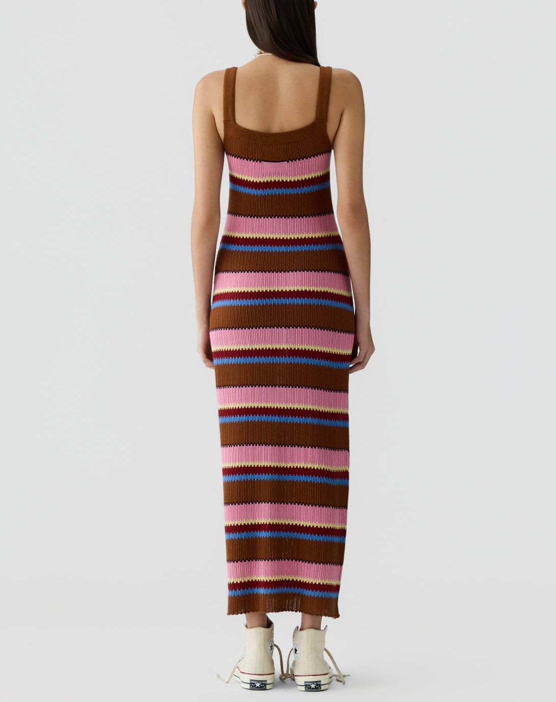 A woman stands facing away, wearing the Guest in Residence Palazzo Dress—a sleeveless, ankle-length silk knit with horizontal stripes in brown, pink, purple, and blue tones—and pairs it with white shoes.