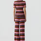 A person stands facing away, wearing the Palazzo Top by Guest in Residence—a vacation-ready linen-silk set with short sleeves and wide-leg pants in pink, brown, purple, and blue horizontal stripes.
