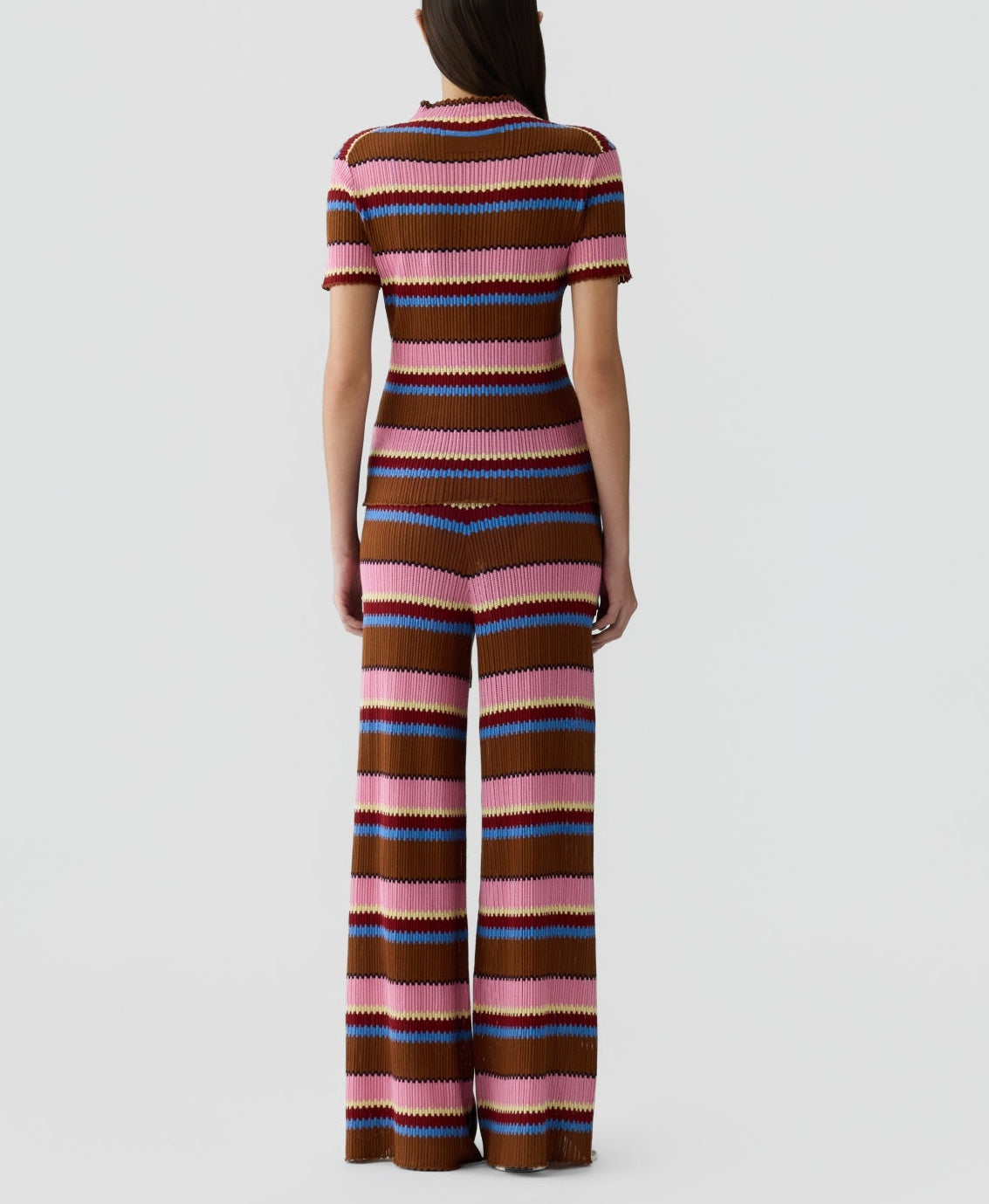 A person stands facing away, wearing the Palazzo Top by Guest in Residence—a vacation-ready linen-silk set with short sleeves and wide-leg pants in pink, brown, purple, and blue horizontal stripes.