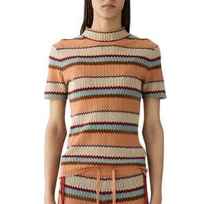 A woman wears the Palazzo Top by Guest in Residence, a breathable knit short-sleeve top with horizontal stripes in peach, beige, brown, mint green, and burgundy—styled here with the matching skirt for a vacation-ready look.