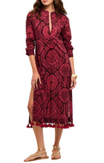 A woman wearing the Figue Paolina Kaftan, a long-sleeve red and black band collar dress with a front slit, tassel hem, textured cotton burnout fabric, paired with gold sandals.