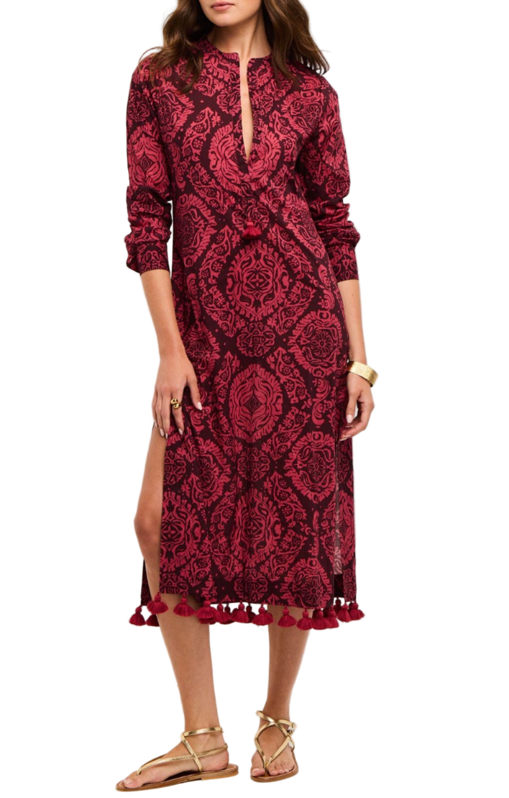 A woman wearing the Figue Paolina Kaftan, a long-sleeve red and black band collar dress with a front slit, tassel hem, textured cotton burnout fabric, paired with gold sandals.