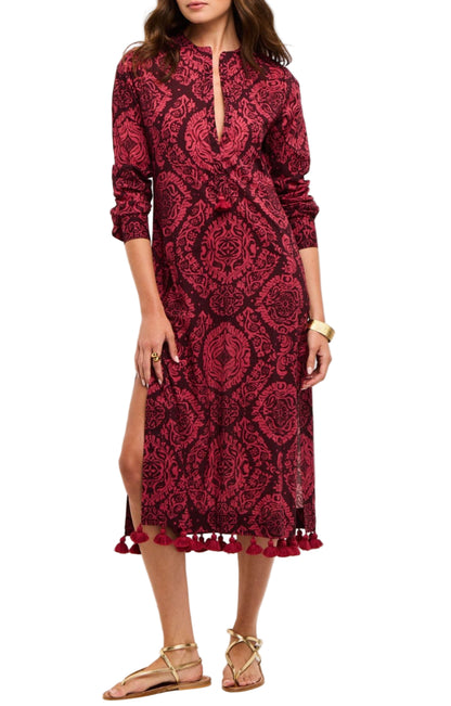 A woman wearing the Figue Paolina Kaftan, a long-sleeve red and black band collar dress with a front slit, tassel hem, textured cotton burnout fabric, paired with gold sandals.