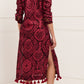 A woman faces away wearing the Figue Paolina Kaftan, a long-sleeved burgundy dress featuring a red damask pattern, band collar, side slit, tassels at the hem, and gold sandals.