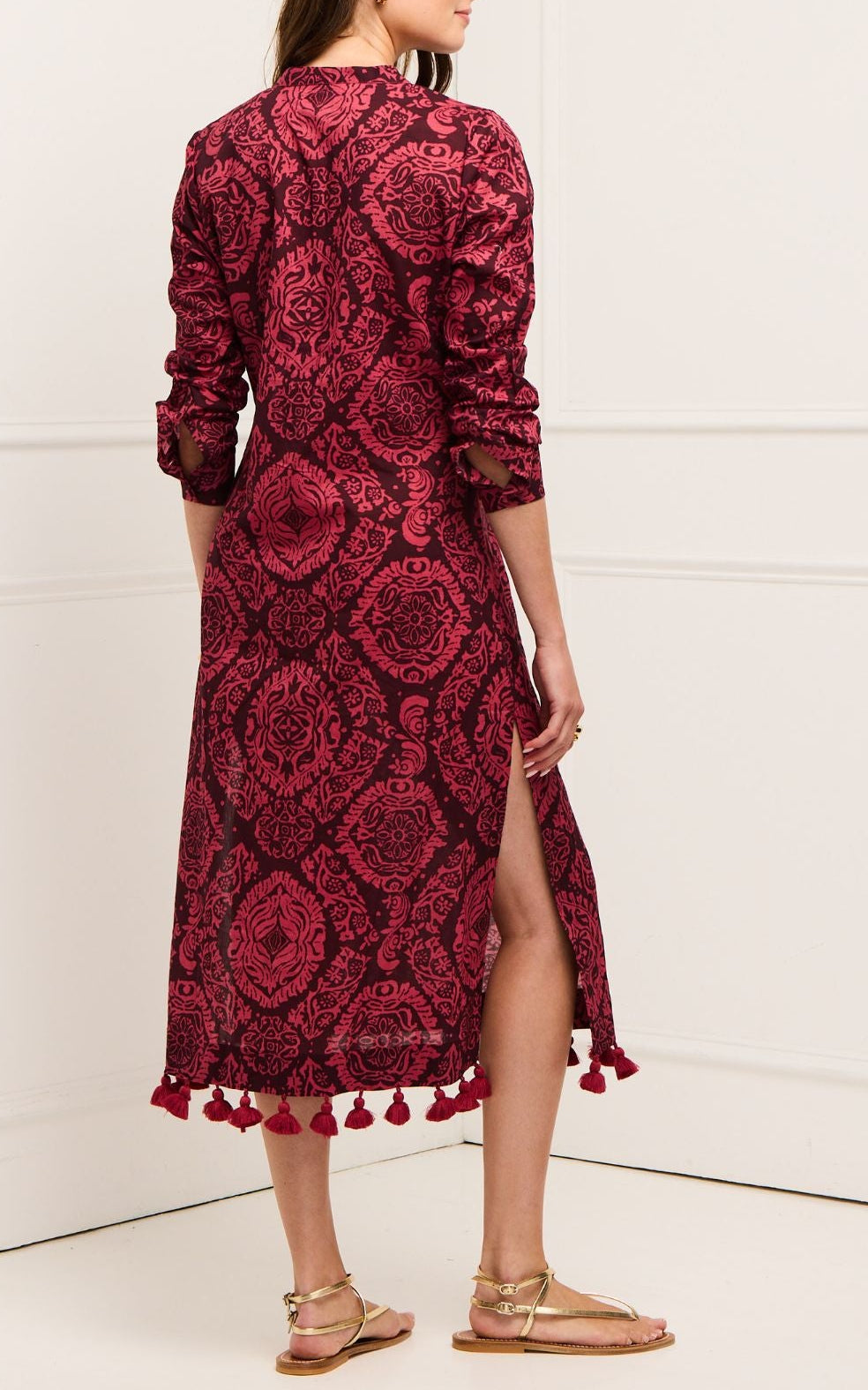 A woman faces away wearing the Figue Paolina Kaftan, a long-sleeved burgundy dress featuring a red damask pattern, band collar, side slit, tassels at the hem, and gold sandals.