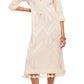 A woman wears the Figue Paolina Kaftan, a light beige long sleeve dress with a geometric pattern, band collar, tassel hem, and custom embroidery. She pairs it with gold sandals and rests one hand on her hip.
