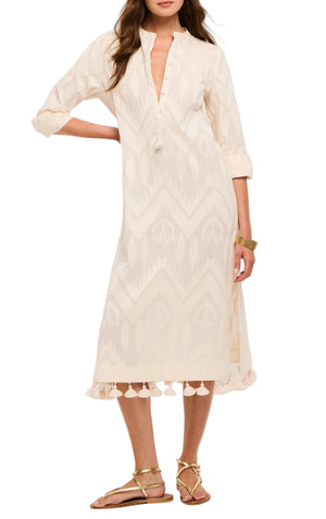 A woman wears the Figue Paolina Kaftan, a light beige long sleeve dress with a geometric pattern, band collar, tassel hem, and custom embroidery. She pairs it with gold sandals and rests one hand on her hip.