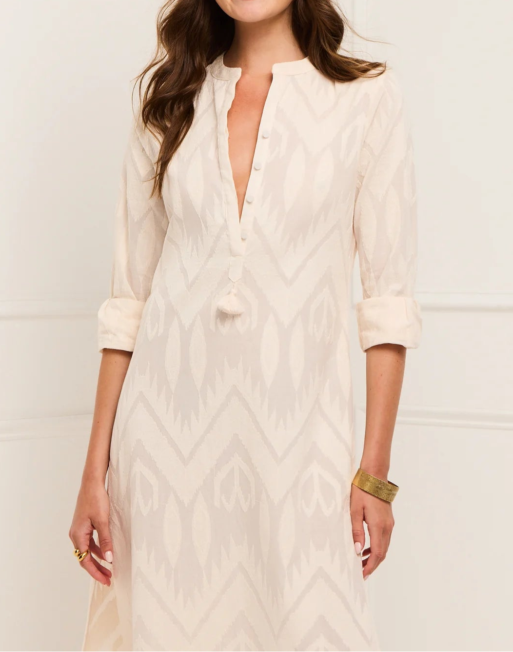 A woman wears the Figue Paolina Kaftan—a cream-colored, patterned, long-sleeve band collar dress with rolled-up sleeves and a partial button-down front—paired with a gold bracelet.