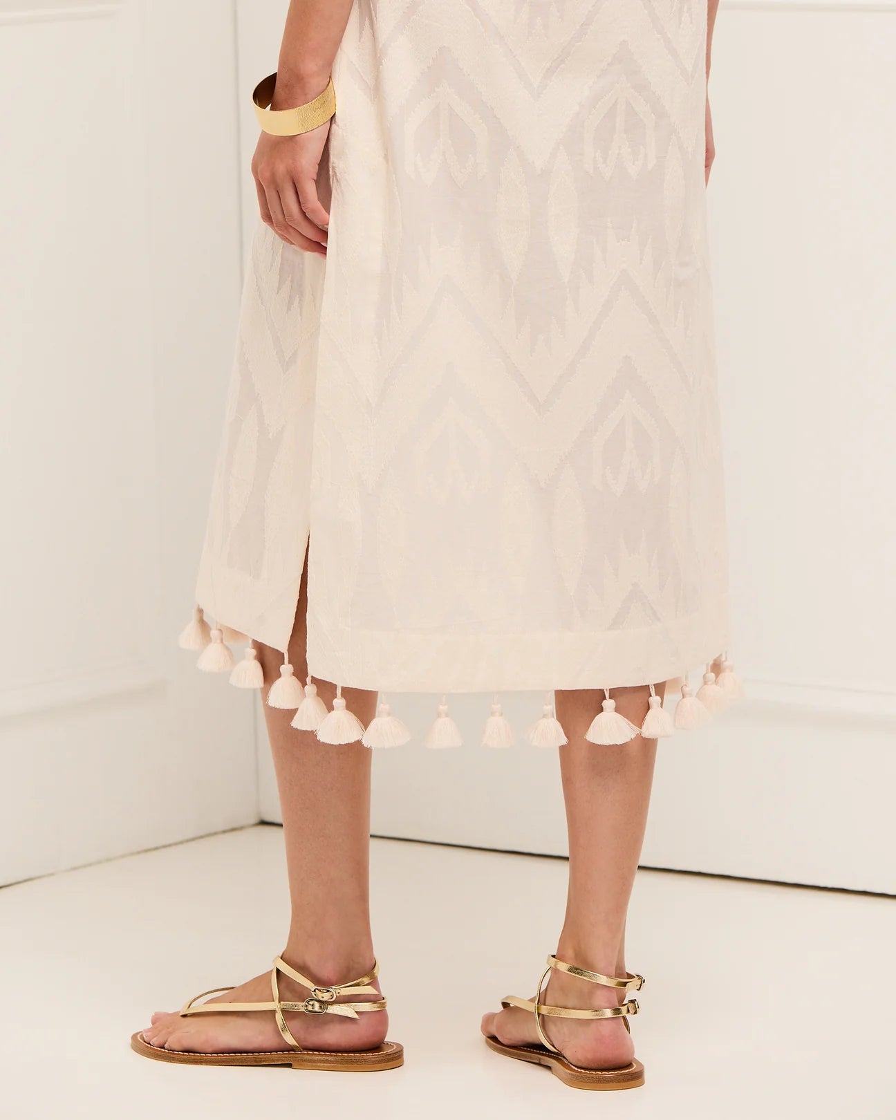 Indoors, a person wears the Figue Paolina Kaftan—a light-patterned, cotton burnout midi dress with a band collar and tassel-trimmed hem—paired with gold bracelets and tan strappy sandals for a textured, elegant look.