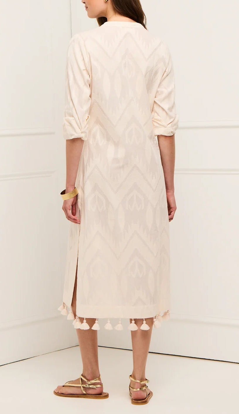 A woman faces away from the camera, wearing the Figue Paolina Kaftan—a light, knee-length cotton burnout dress with rolled sleeves and tassels at the hem—paired with flat sandals.